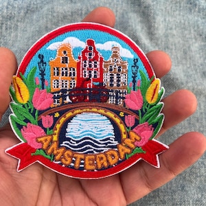 May include: Embroidered patch featuring a colorful Amsterdam scene. The design includes canal houses, a bridge, tulips, and the word "AMSTERDAM" in orange. The patch has a red border and is held in a hand.