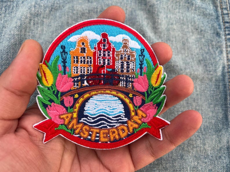 May include: Embroidered patch featuring a colorful Amsterdam scene. The design includes canal houses, a bridge, tulips, and the word "AMSTERDAM" in orange. The patch has a red border and is held in a hand.