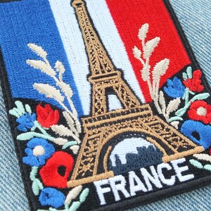 Peut inclure: Écusson brodé représentant la Tour Eiffel, le drapeau français et des motifs floraux. Le mot "FRANCE" est brodé en blanc. L'écusson est rectangulaire avec un bord noir, sur un fond en jean.