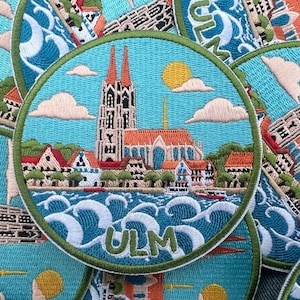 Könnte beinhalten: Gestickter Aufnäher mit einer detaillierten Illustration von Ulm, Deutschland. Das Design zeigt das Ulmer Münster, Gebäude, Wasser und das Wort "ULM". Der Aufnäher hat einen türkisfarbenen Hintergrund mit einem grünen Rand.