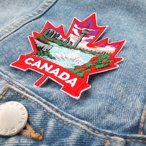 Könnte beinhalten: Gestickter Kanada-Aufnäher in Form eines Ahornblattes, mit einer Wasserfall-Szene, einem Turm und einer Stadtlandschaft. Der rote Aufnäher mit weißem Text ist auf eine hellblaue Jeansjacke genäht.