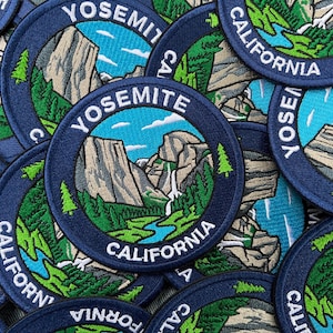 Op de afbeelding: Een ronde geborduurde patch met een landschap van Yosemite, Californië. Het ontwerp bevat beige granieten kliffen, een blauwe rivier, groene dennenbomen en een witte waterval onder een blauwe lucht. De donkerblauwe rand bevat de tekst YOSEMITE CALIFORNIA.