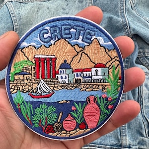 May include: Embroidered patch with a blue border featuring the word "CRETE". The patch depicts a coastal scene with mountains, buildings, a sailboat, and pottery. The color palette includes blue, brown, red, and green.