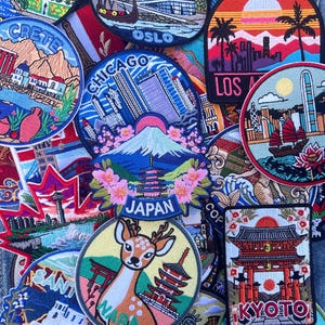 Pode incluir: Uma coleção de patches bordados com destinos de viagem. Os designs incluem paisagens urbanas, marcos e cenas naturais. Os patches mostram lugares como Chicago, Japão e Kyoto, com cores vibrantes e costuras detalhadas. Os patches estão dispostos em um fundo jeans.