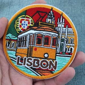 Puede incluir: Parche bordado circular que representa un tranvía amarillo de Lisboa, una bandera portuguesa y el nombre de la ciudad. El parche tiene un borde rojo, verde y naranja. El diseño incluye detalles arquitectónicos y se sostiene en una mano.