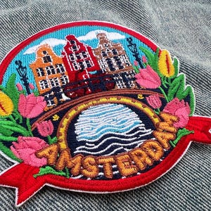 May include: Embroidered patch featuring Amsterdam with colorful canal houses, a bridge, and tulips. The design includes the word "Amsterdam" on a red banner, set against a blue sky and water.
