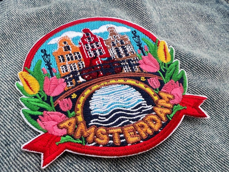 May include: Embroidered patch featuring Amsterdam with colorful canal houses, a bridge, and tulips. The design includes the word "Amsterdam" on a red banner, set against a blue sky and water.