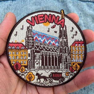 May include: Embroidered patch featuring the word "VIENNA" above a detailed illustration of St. Stephen's Cathedral, with musical notes and a horse-drawn carriage. The patch has a brown border and is held in a hand.