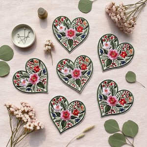 Peut inclure: Écussons brodés en forme de cœur avec des motifs floraux rouges, roses, blancs et bleus. Les écussons sont entourés de feuilles vertes et d'une bordure verte. Des accessoires de couture et des fleurs séchées sont également présents.