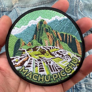 May include: Embroidered patch featuring a detailed depiction of Machu Picchu, with green, brown, and white architectural elements set against a backdrop of green and blue mountains. The patch has a black border and the words "MACHU PICCHU" in green.