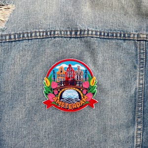 May include: A denim jacket with a colorful embroidered patch. The patch features a cityscape with buildings, a bridge, and the word "AMSTERDAM" in red. The patch is surrounded by flowers and greenery.