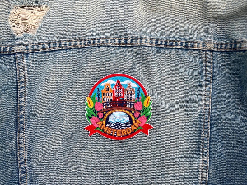 May include: A denim jacket with a colorful embroidered patch. The patch features a cityscape with buildings, a bridge, and the word "AMSTERDAM" in red. The patch is surrounded by flowers and greenery.
