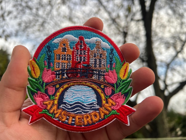 May include: Embroidered patch featuring a colorful depiction of Amsterdam. The design includes canal houses, a bridge, and tulips, with the city's name in a red banner. The patch is surrounded by a blue border.