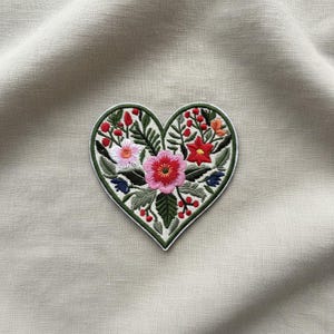 Puede incluir: Parche bordado en forma de corazón con un diseño floral. El parche tiene un borde verde y está relleno de flores rosas, rojas, blancas y azules, hojas verdes y bayas rojas. El parche está sobre un fondo de tela neutra.