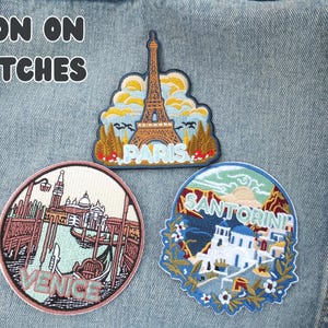 Puede incluir: Tres parches bordados para planchar con temas de viaje. Uno muestra la Torre Eiffel con el texto "PARIS". Otro muestra Venecia con una g&oacute;ndola y el texto "VENICE". El tercero muestra Santorini con el texto "SANTORINI".