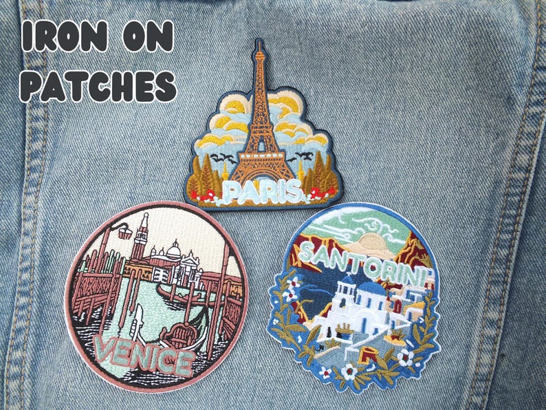 Puede incluir: Tres parches bordados para planchar con temas de viaje. Uno muestra la Torre Eiffel con el texto "PARIS". Otro muestra Venecia con una g&oacute;ndola y el texto "VENICE". El tercero muestra Santorini con el texto "SANTORINI".