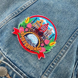 May include: Embroidered patch featuring the word "AMSTERDAM" in red, with a circular design of buildings, water, and flowers. The patch is on a denim jacket, showcasing a vibrant and detailed travel-themed accessory.