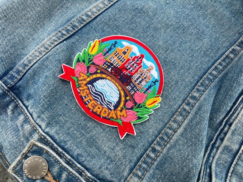 May include: Embroidered patch featuring the word "AMSTERDAM" in red, with a circular design of buildings, water, and flowers. The patch is on a denim jacket, showcasing a vibrant and detailed travel-themed accessory.