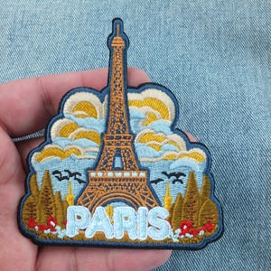 May include: Embroidered patch featuring the Eiffel Tower in Paris, France. The design includes the tower in brown, blue sky with yellow clouds, trees, and the word "PARIS" in light blue. The patch is bordered in dark blue.