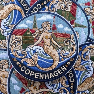 May include: Embroidered patch featuring a mermaid in front of a cityscape with the word "COPENHAGEN". The patch has a blue border and a detailed design with red, blue, and tan colors. The mermaid is the central focus.