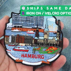 Puede incluir: Parche bordado que representa Hamburgo, Alemania, con edificios, barcos y el nombre de la ciudad en rojo. El parche tiene un fondo azul y un borde negro. El texto en el parche dice "HAMBURG".