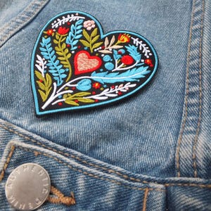 Puede incluir: Un parche bordado en forma de corazón azul con un diseño floral y un corazón rojo en el centro. El parche está en una chaqueta vaquera azul.