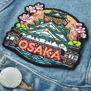 Peut inclure: Écusson brodé représentant le château d'Osaka, des fleurs de cerisier et le mot "OSAKA" en rouge. L'écusson est fixé sur une veste en jean. L'écusson a une bordure noire et est coloré.