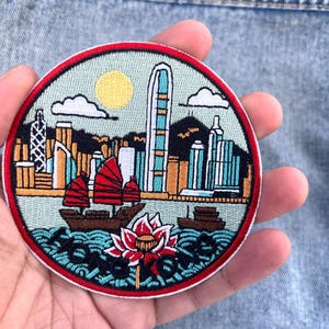 May include: Embroidered round patch featuring a cityscape with a red-sailed boat, a lotus flower, and the text "Hong Kong". The patch has a red border, a yellow sun, and blue water. The buildings are in shades of brown, blue, and white.