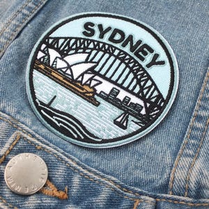 May include: A blue and white embroidered patch featuring the word "Sydney" and an image of the Sydney Opera House and Harbour Bridge.