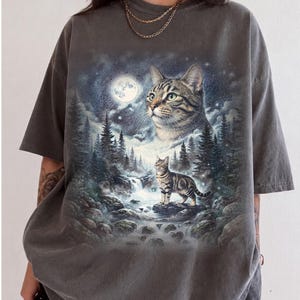 May include: A charcoal gray short-sleeve t-shirt featuring a graphic print of a tabby cat in a forest landscape. The design includes a large cat head in the sky, a smaller cat standing on a rock by a waterfall, a full moon, and pine trees.