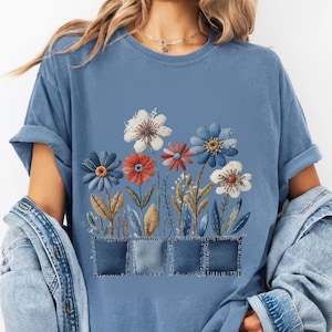 Puede incluir: Camiseta azul claro con un diseño floral bordado. El diseño incluye flores blancas, rojas y azules, con hojas verdes y marrones. Las flores están sobre tres parches de mezclilla.