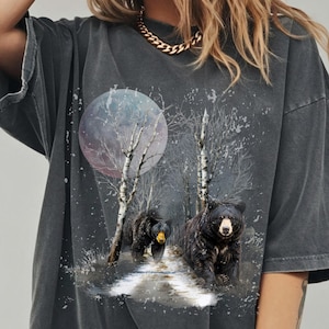 Puede incluir: Camiseta gris oscuro con una escena de bosque con dos osos negros caminando por un sendero. Una luna llena y abedules en el fondo. La camiseta tiene un aspecto vintage.
