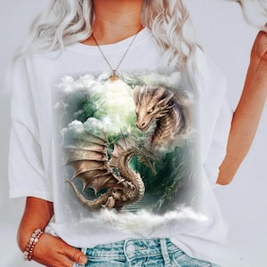 May include: White t-shirt featuring a detailed fantasy illustration of two dragons amidst clouds and a mountainous backdrop. One dragon is in flight, wings spread, while the other is a close-up of a dragon's head. The artwork is rendered in shades of brown, green, and white.
