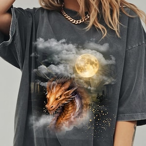 May include: Dark gray t-shirt featuring a detailed illustration of a dragon with orange and brown scales, set against a backdrop of a full moon, clouds, and a spooky landscape. The dragon's head is the focal point.