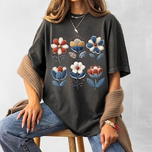 Puede incluir: Camiseta gris oscuro con un diseño floral en rojo, blanco y azul. Las flores están dispuestas en dos filas de tres, con tallos y hojas azules. La camiseta tiene un corte holgado.