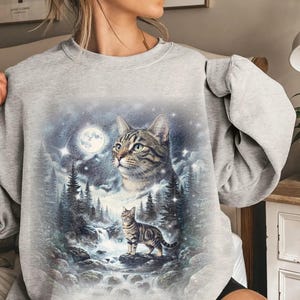 May include: A light gray crewneck sweatshirt featuring a graphic print of a tabby cat in a forest landscape. The design includes a large cat head above a smaller cat standing on a rock near a waterfall, surrounded by pine trees and a full moon.