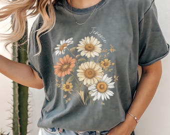 Op de afbeelding: Een grijze t-shirt met een bloemenontwerp van madeliefjes in wit, geel en oranje. Het woord "Jeneece" staat boven de bloemen. De t-shirt heeft korte mouwen en een relaxte pasvorm. De persoon draagt een spijkerbroek.