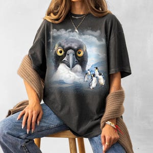 Puede incluir: Camiseta gris oscuro con un diseño de un ave con ojos amarillos y pecho blanco, con un fondo de pingüinos y un paisaje nevado. La camiseta es de corte holgado.