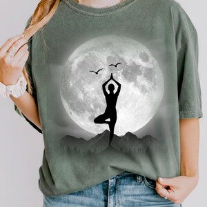 以下が含まれることがあります： セージグリーンのTシャツ。満月の前でヨガのポーズをとる人の黒いシルエットが描かれています。2羽の鳥が月の前を飛んでいます。デザインは山脈を背景にしています。