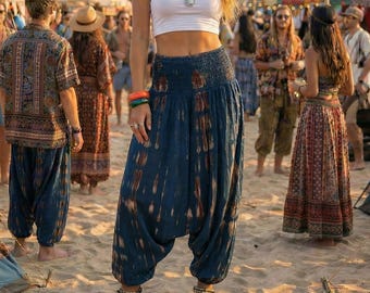 May include: A woman in a white tube top and blue harem pants stands on a sandy beach. She wears multiple bracelets, necklaces, and anklets. The background includes tents, flags, and a carousel, suggesting a festival or market setting.
