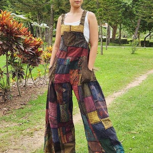 Pode incluir: Um macacão de patchwork com uma variedade de quadrados de tecido em tons de vermelho, amarelo, azul, verde e castanho. A peça tem pernas largas, alças grossas, um bolso no peito e dois bolsos laterais, usado sobre uma camisola sem mangas branca.