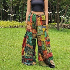 以下が含まれることがあります： 黒いタンクトップとパッチワークのパンツを履いた女性。パンツは緑、茶色、赤、青など、さまざまな生地と色で作られています。女性は木々の背景に草が生えている場所に立っています。