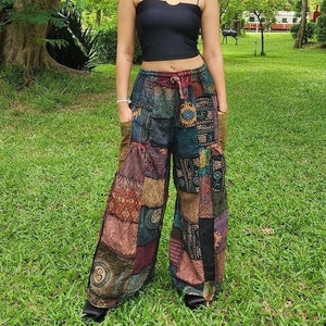 Pode incluir: Uma mulher usando um top preto e calças patchwork. As calças são feitas de vários padrões de tecido em tons de marrom, verde e roxo. A mulher está em pé em um gramado com árvores ao fundo.