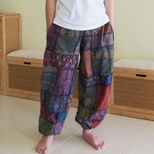 以下が含まれることがあります： 白い半袖Tシャツとパッチワークパンツを履いた人。パンツは紫、青、緑など、さまざまな色と模様が特徴です。絵画と木製の家具の前に立っています。