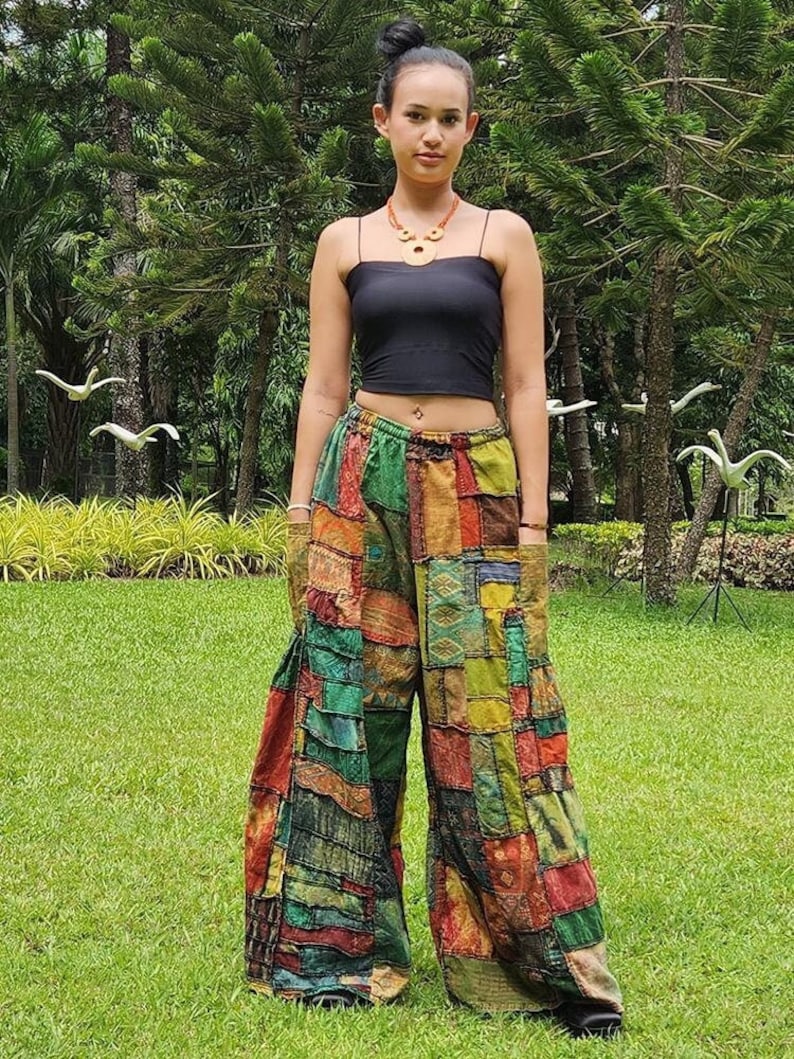 May include: A woman wearing a black tank top and patchwork pants. The pants are made of different fabrics and colors, including green, brown, red, and blue. The woman is standing in a grassy area with trees in the background.