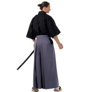 May include: A man wearing a black kimono and grey hakama trousers. He is holding a black sword in his right hand.