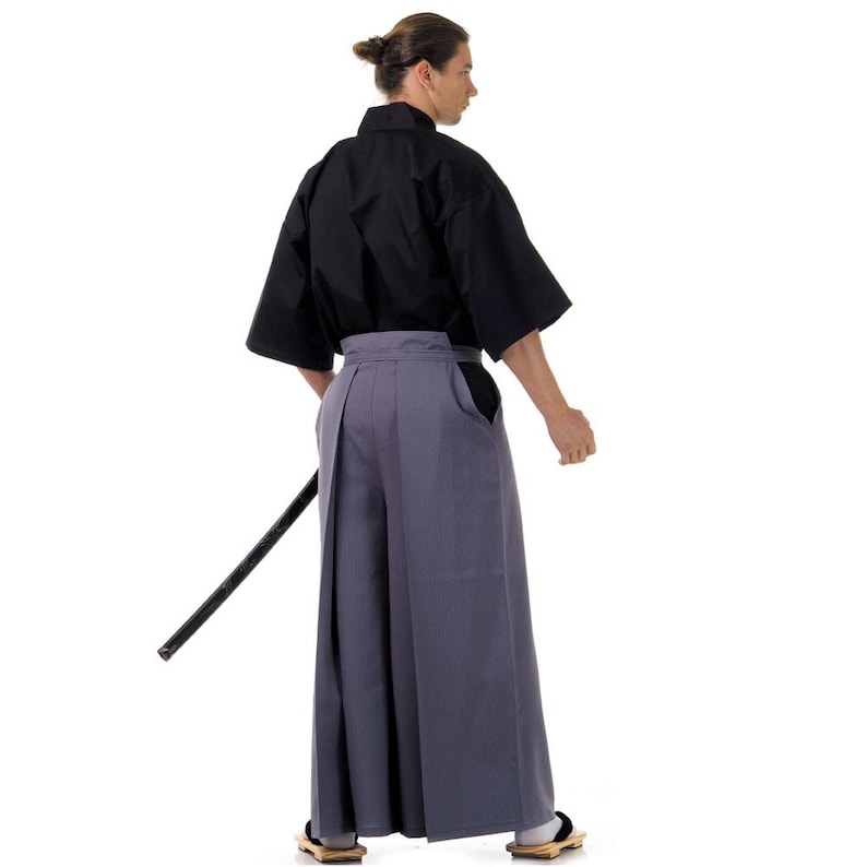 May include: A man wearing a black kimono and grey hakama trousers. He is holding a black sword in his right hand.
