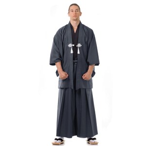 May include: A man wearing a dark blue traditional Japanese robe with white accents and a black sash. He is standing with his hands in his pockets.
