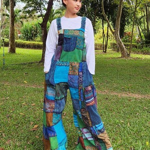 May include: A woman wearing a patchwork jumpsuit with a variety of colors and patterns, including blue, green, brown, and red. The jumpsuit has a wide leg and a relaxed fit. The woman is standing in a grassy area with trees in the background.