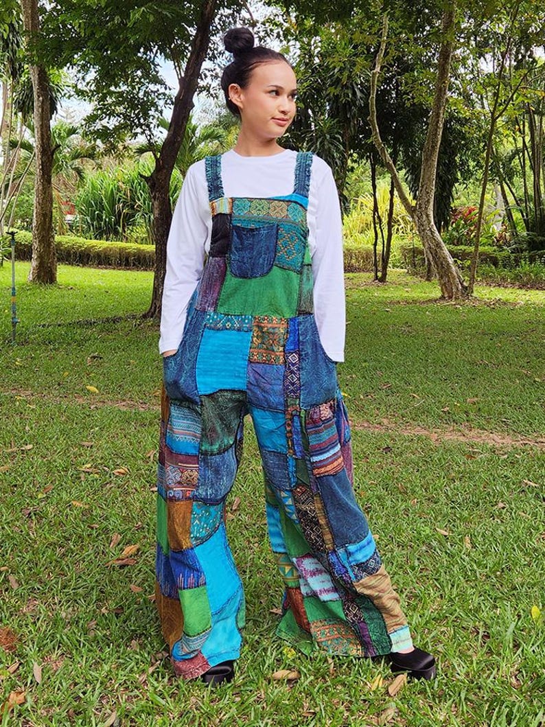 May include: A woman wearing a patchwork jumpsuit with a variety of colors and patterns, including blue, green, brown, and red. The jumpsuit has a wide leg and a relaxed fit. The woman is standing in a grassy area with trees in the background.